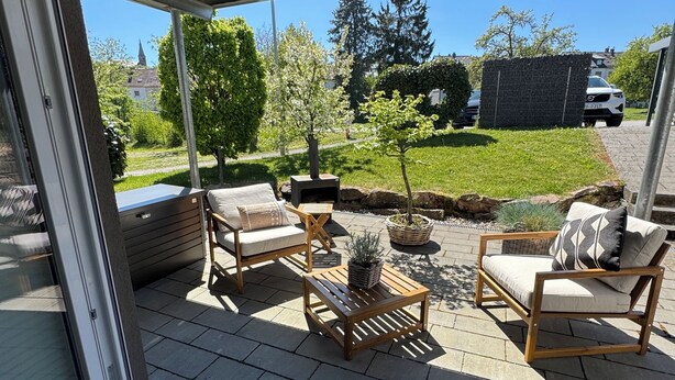 A patio with outdoor furniture, a small table, and a view of a green landscape with trees and a distant building. - Modern vacation apartment with feel-good terrace in Beerfelden (Oberzent)