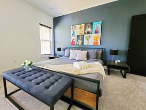 Een moderne slaapkamer met een groot bed, een bankje, twee nachtkastjes met lampen, een raam met zonwering en een wand met bloemkunstwerk.