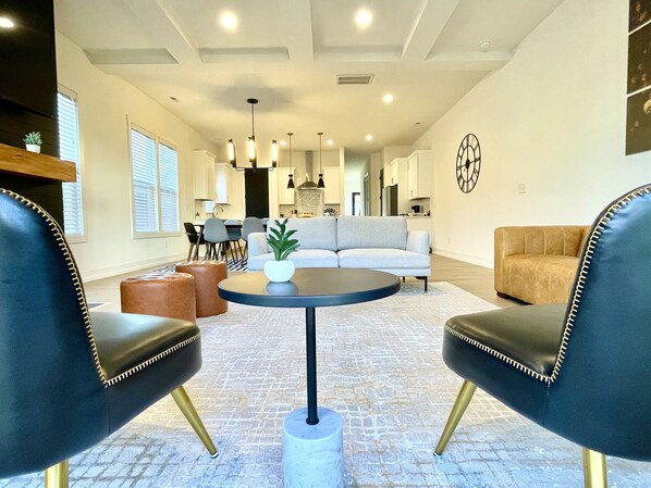 Een moderne woonkamer met een bank, twee lederen fauteuils, een salontafel en een eethoek op de achtergrond.
