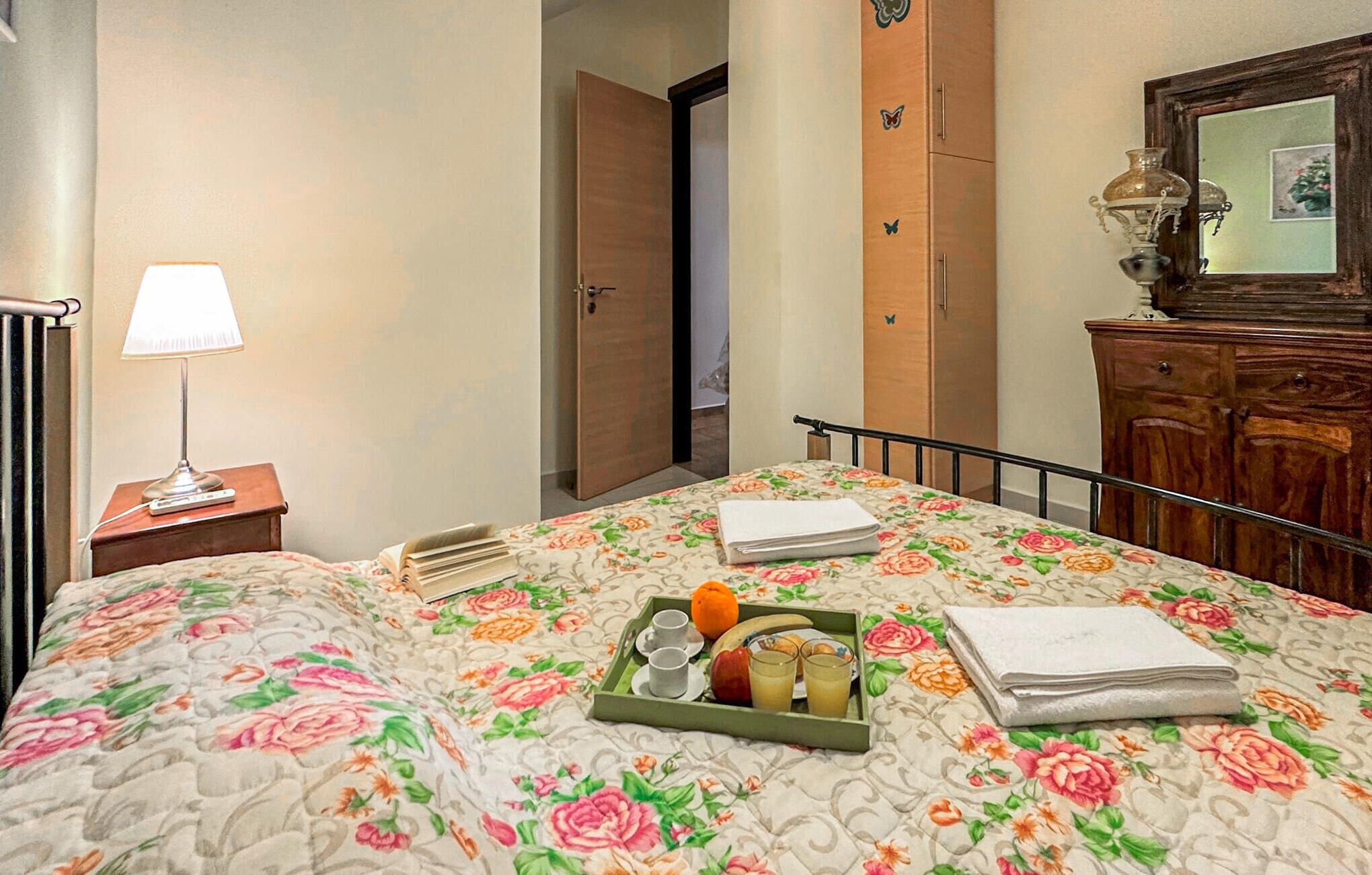 Hotelkamer met een bloemenbedekker, nachtkastje met lamp, dienblad met ontbijtartikelen en een houten dressoir met spiegel.