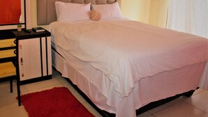 A neatly made bed with a headboard, a bedside table with a lamp, and a red rug on the floor.