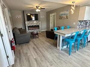Een woonkamer met een open haard, een plafondventilator, een bank en een eethoek met stoelen.