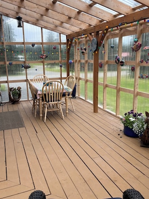 En overdækket terrasse med trægulv, et træbord og stole, samt udsigt gennem store vinduer.