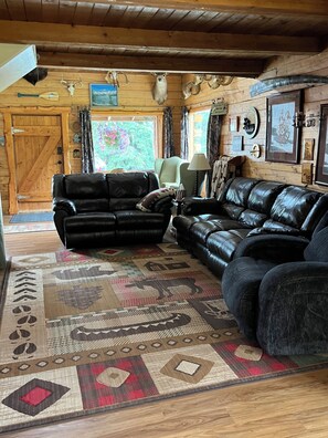 A log cabin interior with two black leather sofas, a patterned rug, wooden walls, and mounted animal skulls.