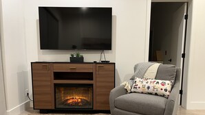 A modern living room with a fireplace, a grey armchair, and a flat-screen TV mounted above the fireplace.