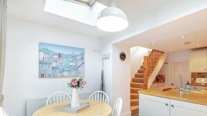Un coin repas avec une table ronde, des chaises blanches, un vase de fleurs, un tableau représentant une ville côtière, et une cuisine avec des meubles blancs et un comptoir en bois.