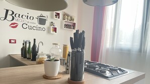 Eine Küchenarbeitsplatte mit verschiedenen Gegenständen, darunter Flaschen, ein Topfpflanze und Besteck. An der Wand hängt ein Schild mit der Aufschrift 'Bacio MEGLIO di come Cucino'.