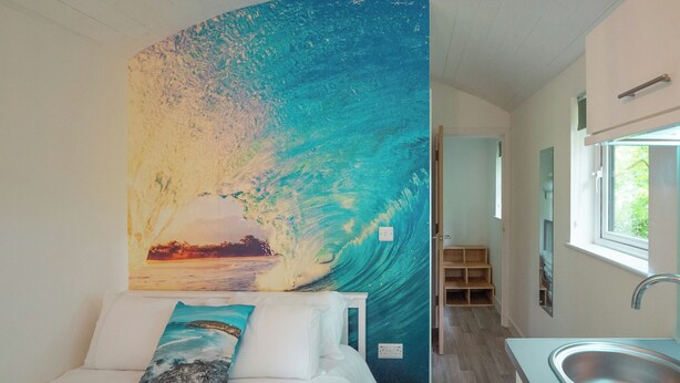 Een slaapkamer met een bed, een kussen, een muurschildering van een golf, een badkamer met een wastafel, en een raam.