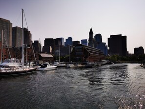 A waterfront cityscape with a prominent clock tower, a river with boats, and modern buildings. - Location+Comfort+Convenience (Boston)