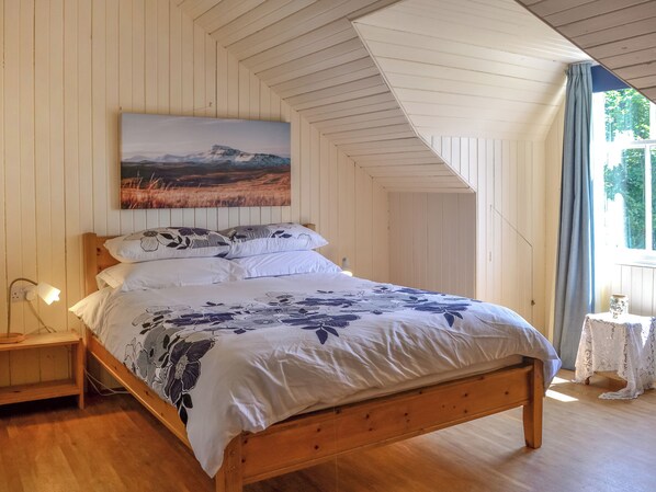 A bedroom with a wooden bed, white walls, a painting above the bed, a nightstand with a lamp, and a window with curtains.