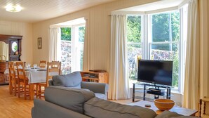 A living room with a sofa, a dining table with chairs, a television, and large windows with curtains. - 3 bedroom accommodation in Rockcliffe, near Dalbeattie (Rockcliffe, near Dalbeattie)