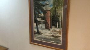 A framed painting of a lighthouse, a brick building, and trees on a wall. - Nautical theme retreat located in the Ivory Mansion  (L'Anse)