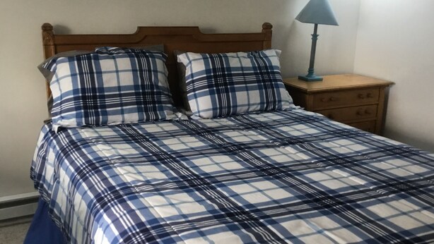 A bed with a plaid bedspread and two pillows, a wooden headboard, a nightstand with a lamp, and a wooden dresser. - Nautical theme retreat located in the Ivory Mansion  (L'Anse)