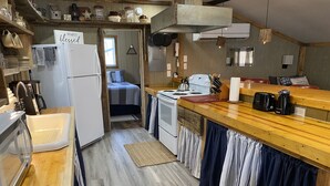 Cocina compacta con refrigerador, estufa y cafetera. Se ve una cama al fondo.