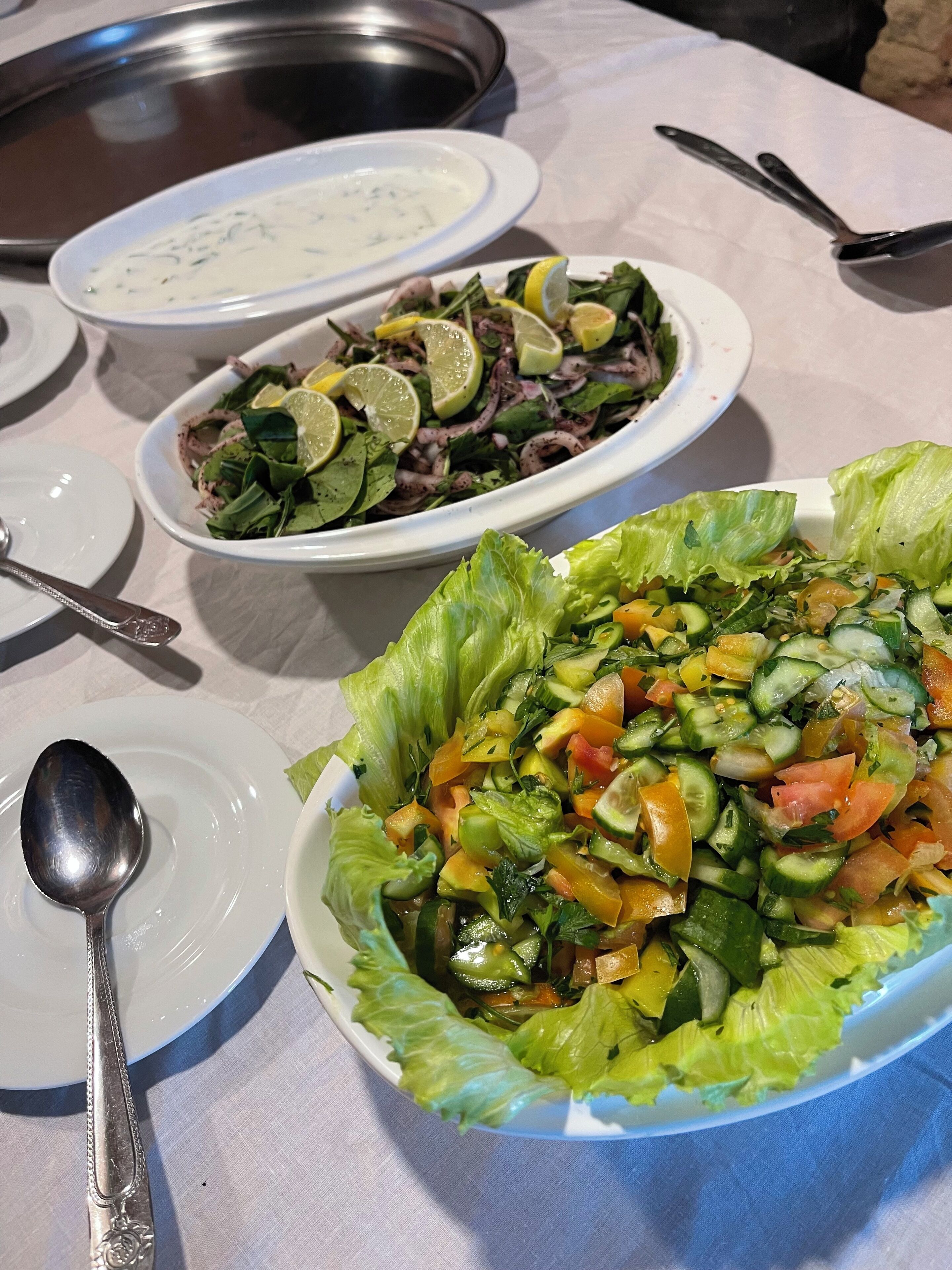 Una mesa con diversos platos, incluyendo una ensalada con pepinos, tomates y lechuga, un tazón de sopa y un plato con otra ensalada.