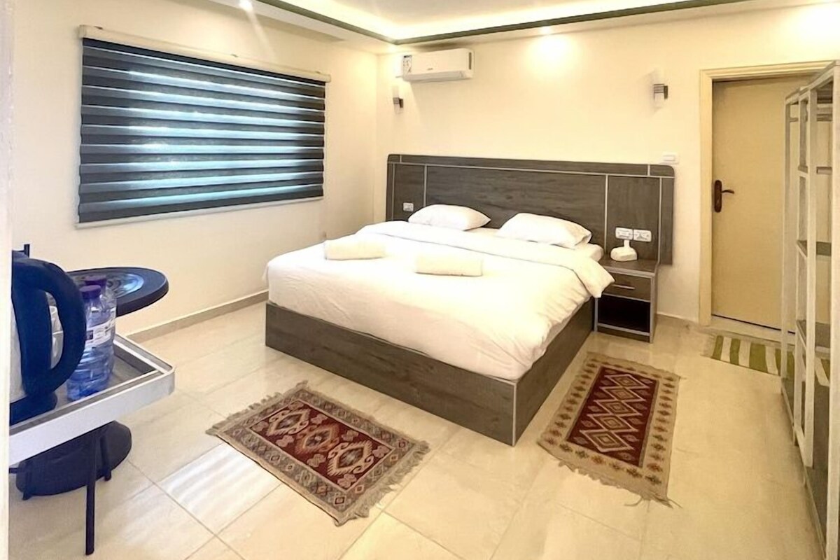 A modern hotel room with a large bed, bedside tables, a water dispenser, and a view of the outside.