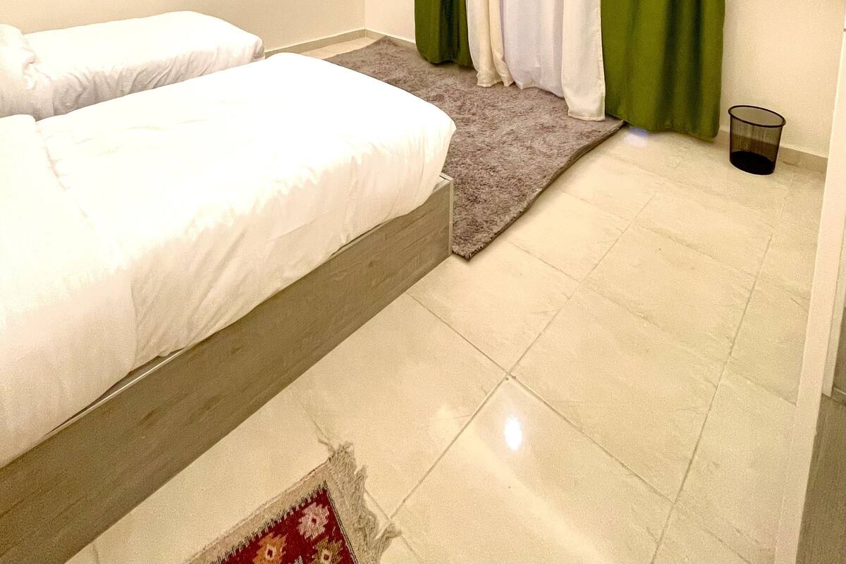 Quarto de hotel com duas camas, um tapete pequeno, uma lixeira e uma janela com cortinas.