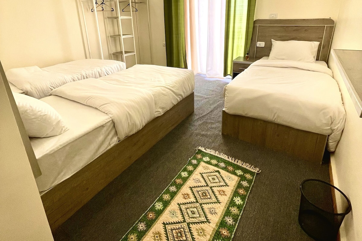 Quarto de hotel com duas camas de solteiro, um guarda-roupa, uma mesinha pequena e um tapete verde estampado.