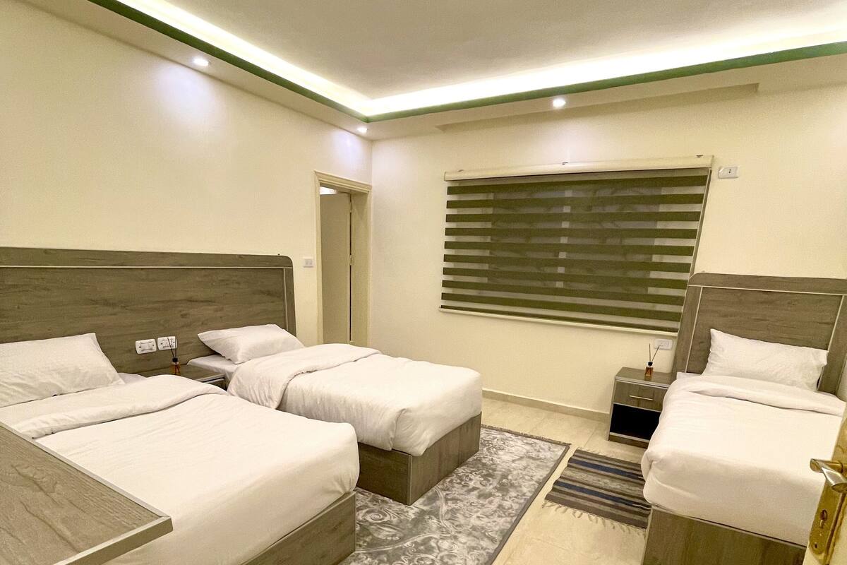 Quarto de hotel moderno com duas camas, uma mesa pequena e uma janela com persianas.