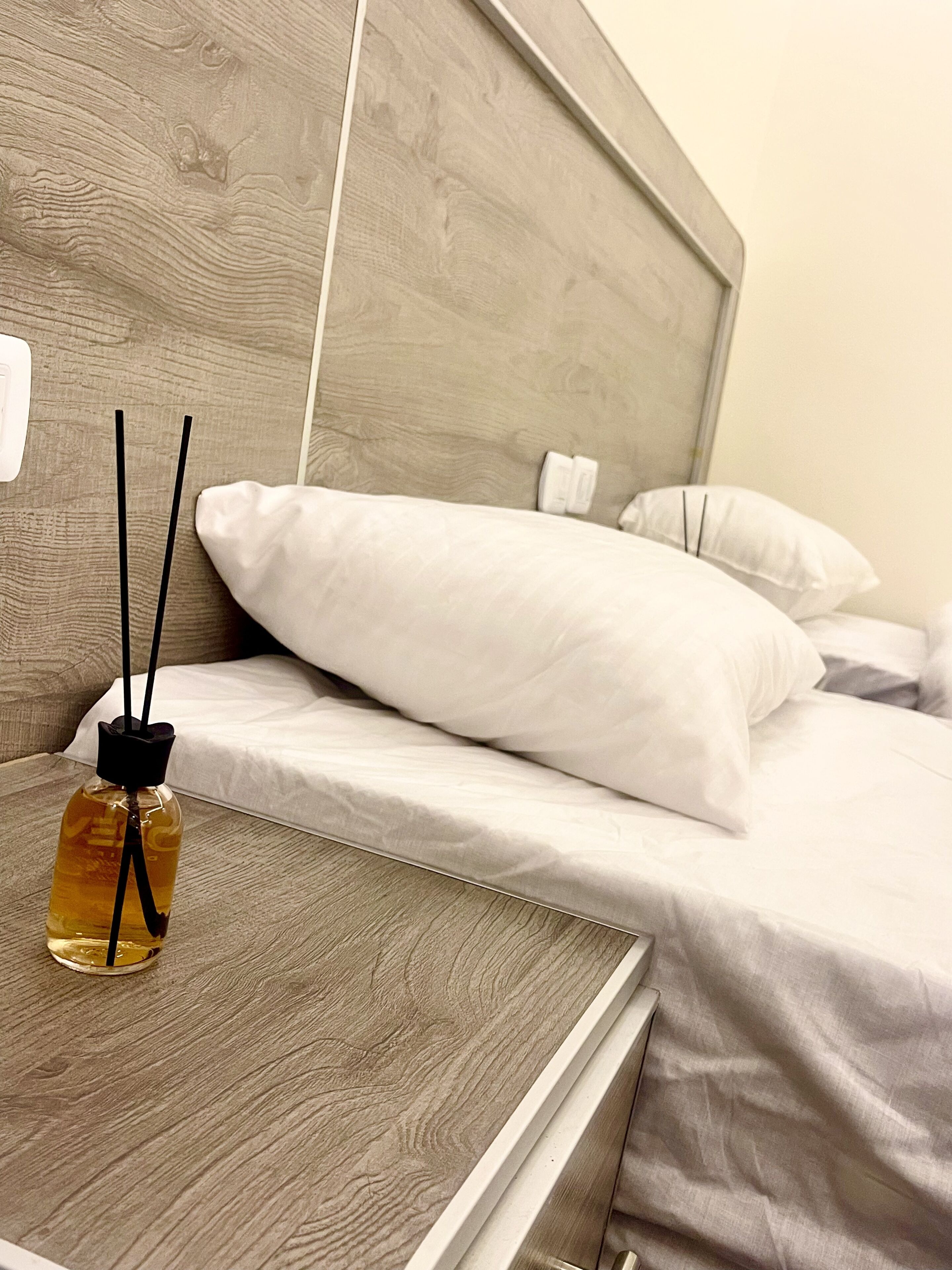Una cama bien tendida con sábanas blancas y un cabecero de madera. En la mesita de noche hay una botella y dos palitos negros.