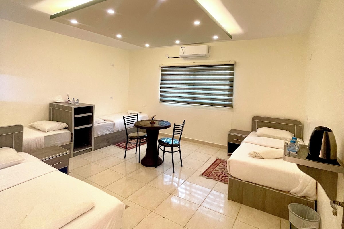 Quarto de hotel com duas camas, uma pequena mesa de jantar, cadeiras, uma máquina de café e uma lixeira.
