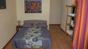 Un dormitorio con una cama, almohadas, una mesita de noche, una lámpara, un cuadro de colores en la pared y un estante de madera con toallas.