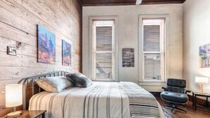 A bedroom with a wooden wall, a bed with striped bedding, two windows with blinds, a black chair, and a small table with a lamp. - Satchmo: Dwntwn Bryan-Main Street, 2nd floor, King Beds (Bryan)