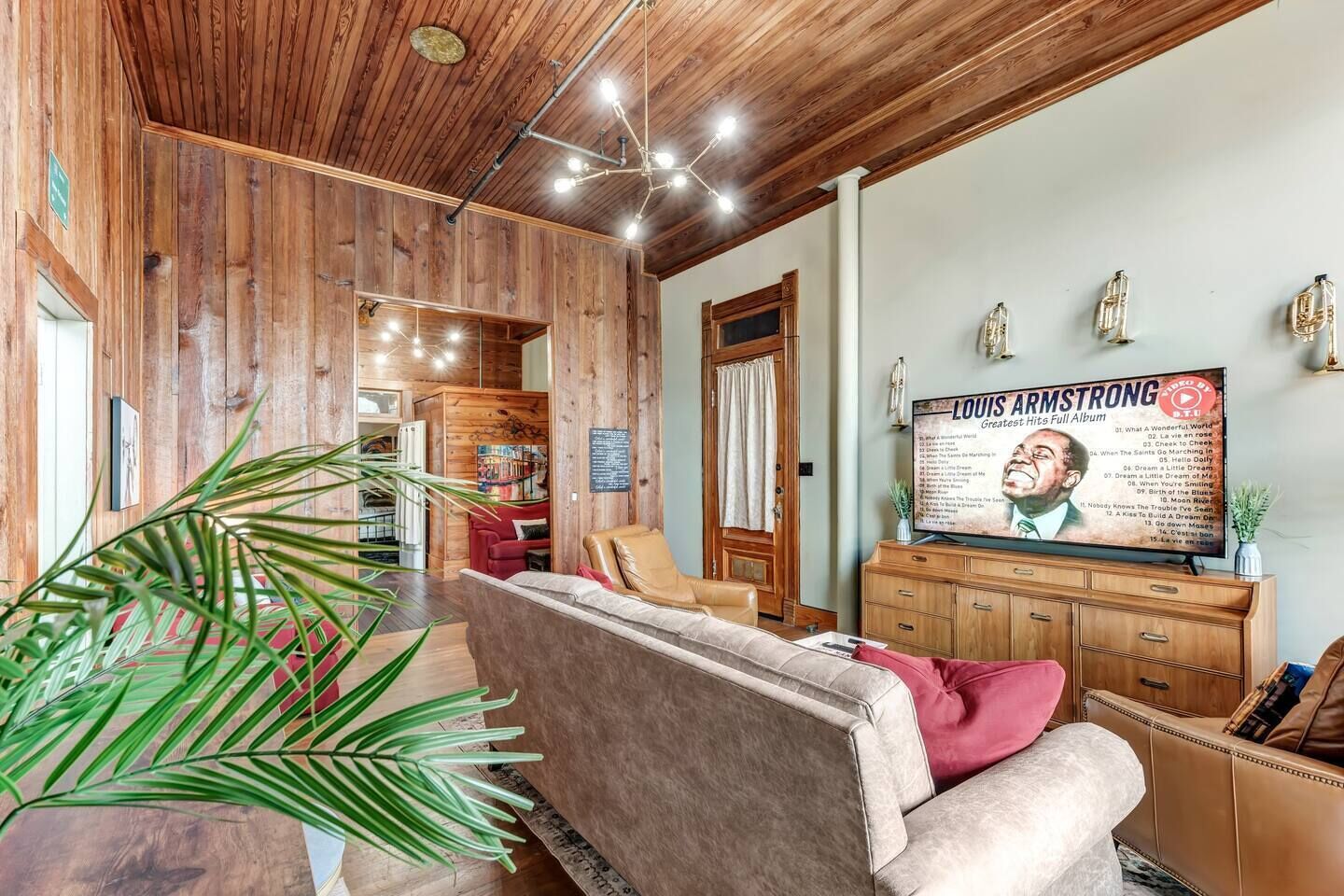 A cozy living room with a wooden ceiling, a large flat-screen TV displaying a Louis Armstrong album, a red sofa, and a wooden cabinet.