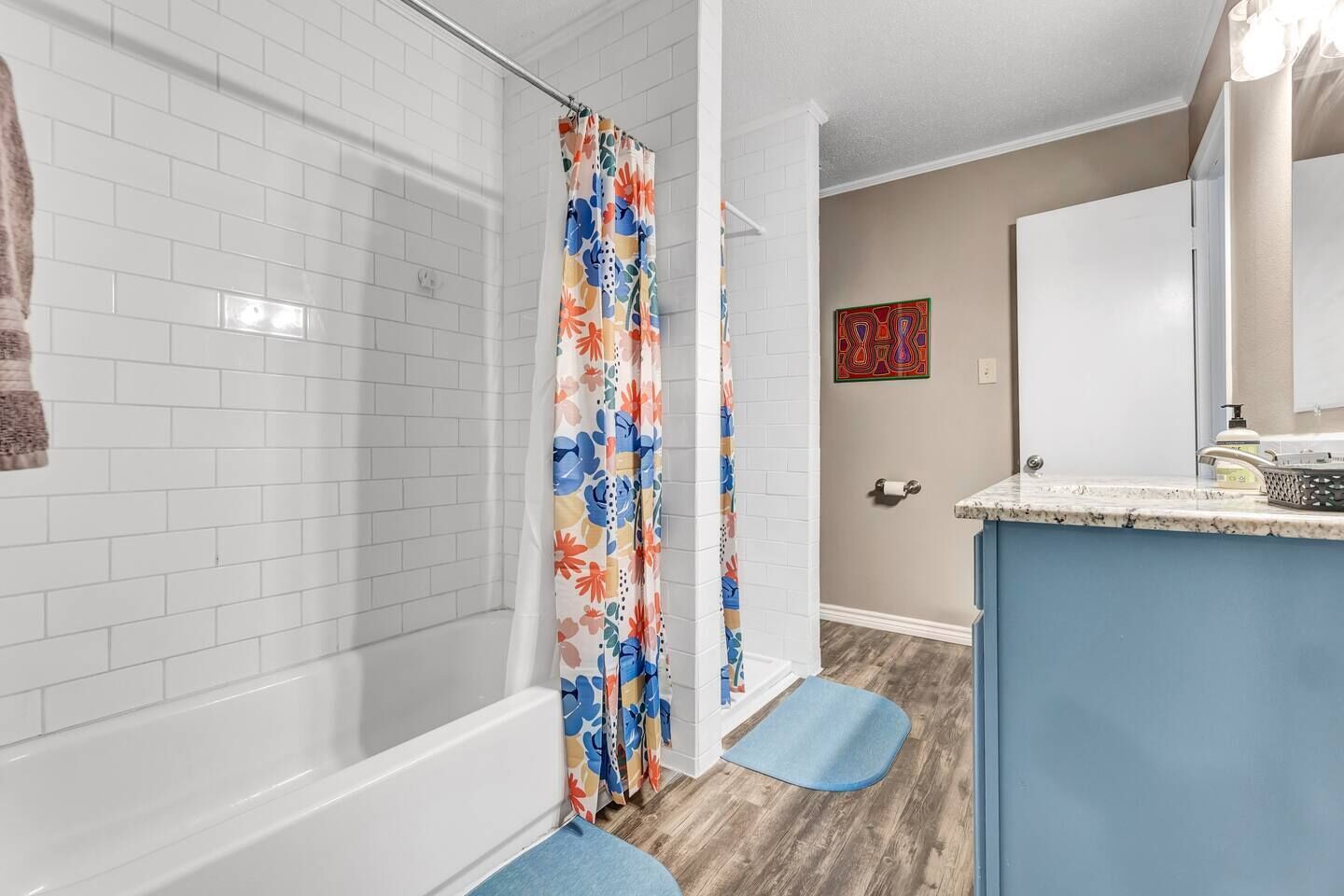 A bathroom with white tiled walls, a shower with a floral curtain, a blue mat, and a sink with a soap dispenser.