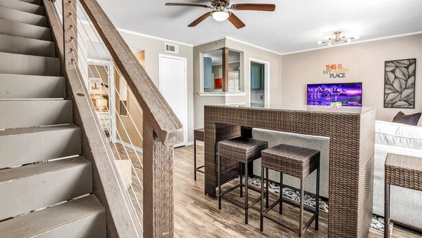 A modern living room with a staircase, a ceiling fan, a bar area with two stools, and a TV on the wall. - St Simons: 3 Bedroom, Walk to A&M, King Beds (Bryan)