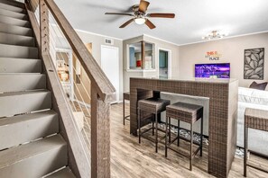 A modern living room with a staircase, a ceiling fan, a bar area with two stools, and a TV on the wall. - St Simons: 3 Bedroom, Walk to A&M, King Beds (Bryan)