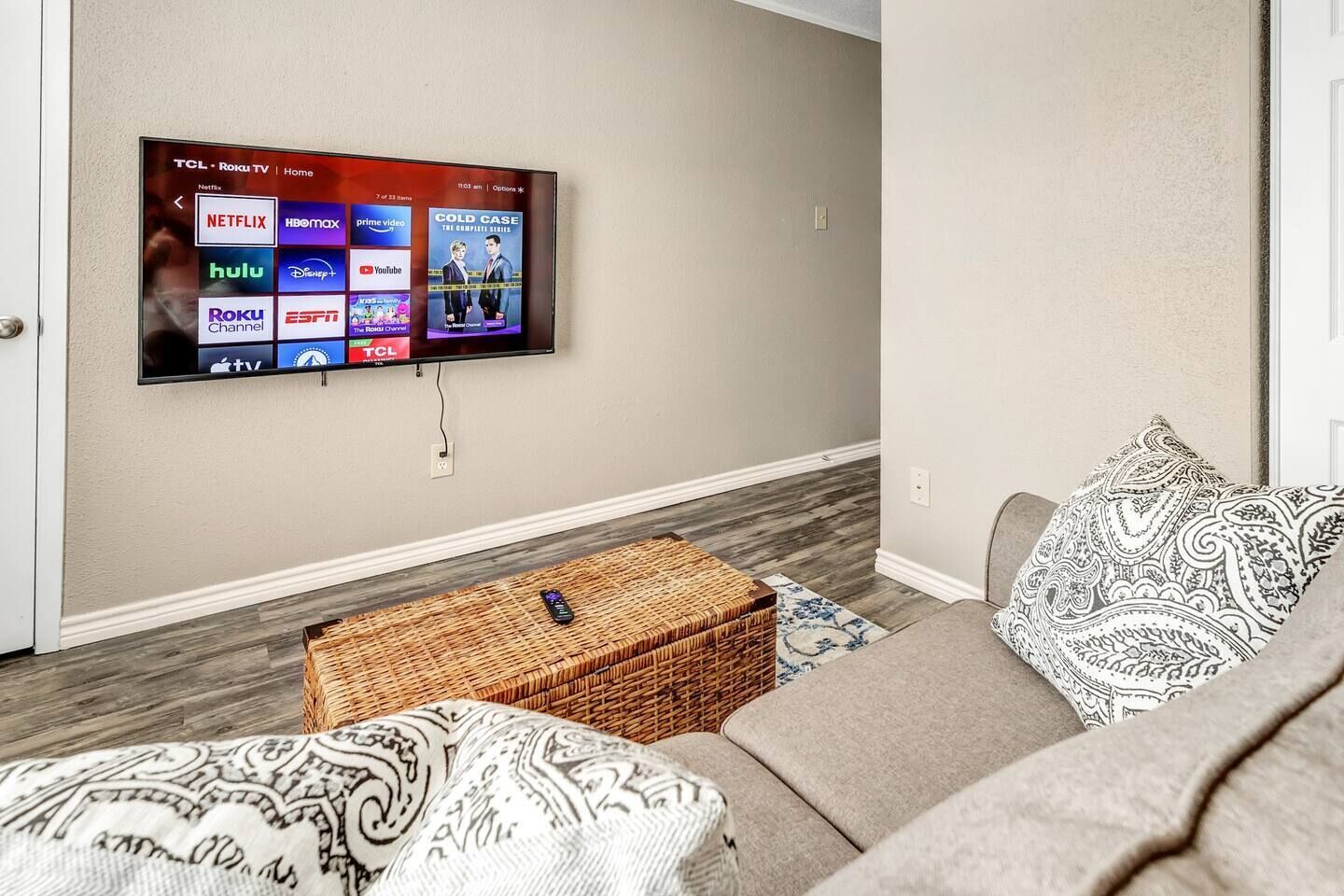 A living room with a flat-screen TV mounted on the wall displaying various streaming services. There is a woven basket on the floor and a patterned throw pillow on the couch.