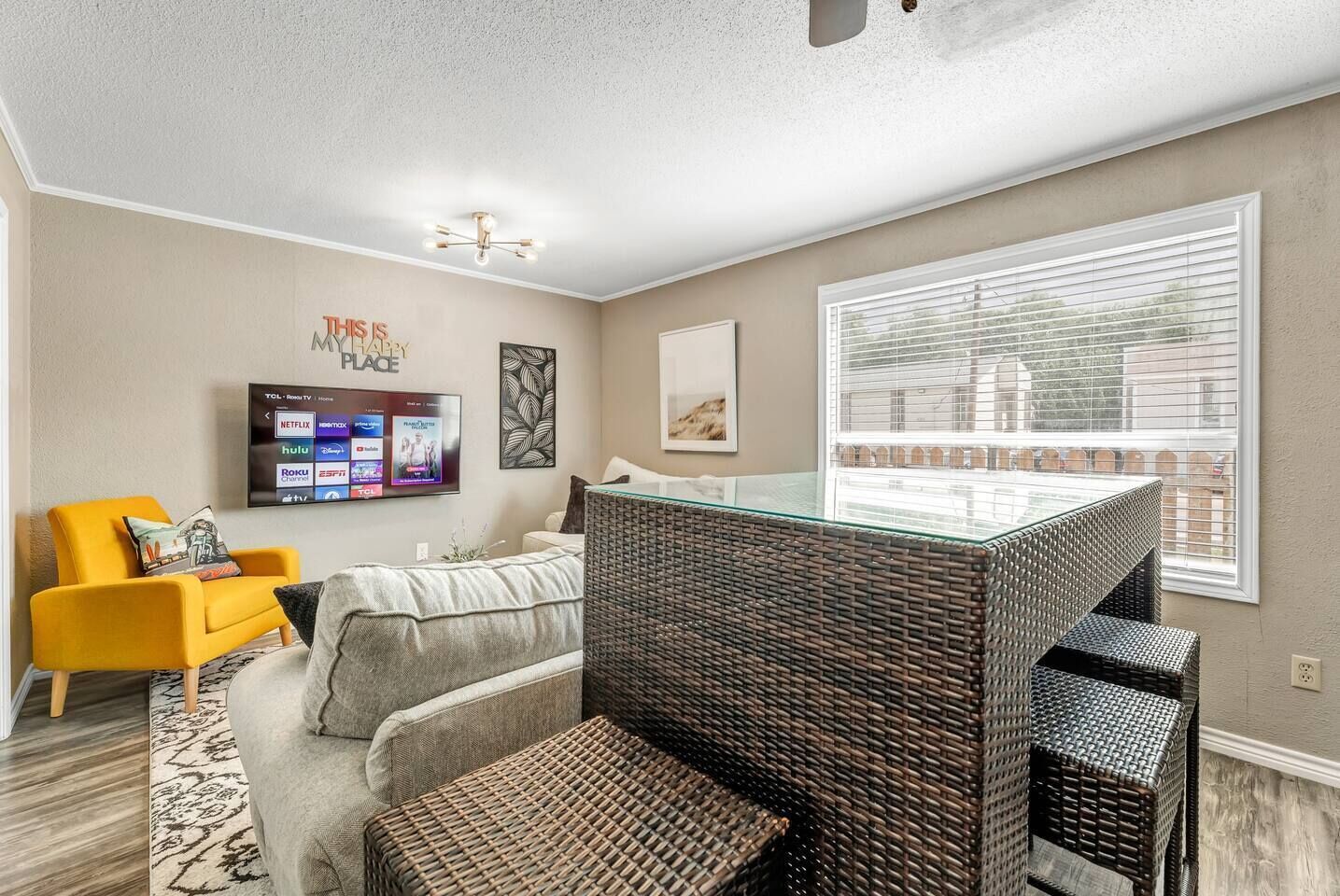 A modern living room with a flat-screen TV displaying streaming services, a comfortable sofa, a yellow armchair, a glass-top coffee table, and a large window with blinds.