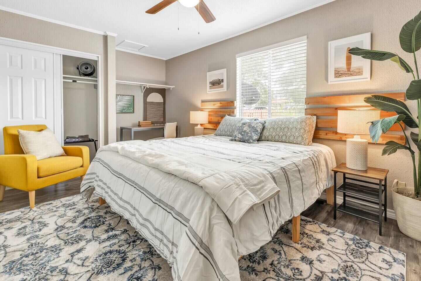 A bedroom with a bed, a yellow armchair, a desk, a window with blinds, and a plant.