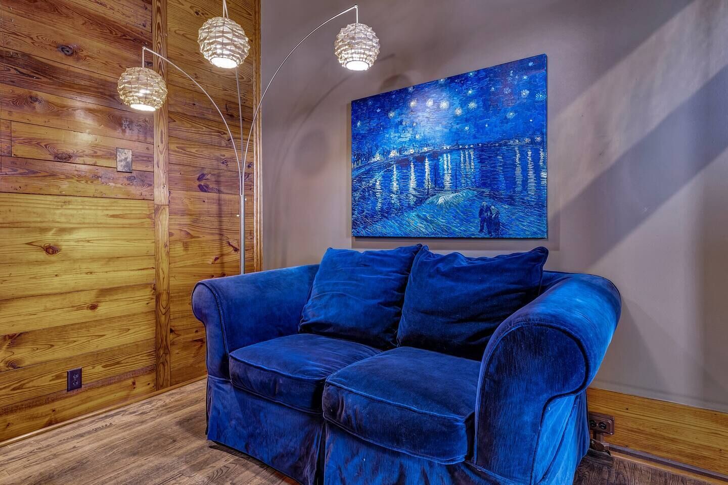 A room with a blue velvet sofa, wooden panel walls, and a painting of a night scene.