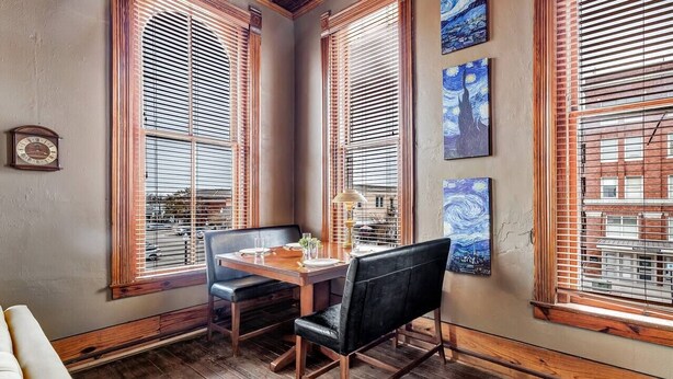 A dining area with a wooden table and chairs, large windows with blinds, and artwork on the walls. - Starry Night: Dwntwn Bryan on Main St, 2nd Flr, King Bed (Bryan)
