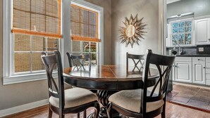 Un coin repas avec une table ronde et quatre chaises, de grandes fenêtres équipées de stores, et une cuisine visible à travers une porte ouverte.