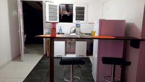 A compact kitchen with a refrigerator, a sink, and a countertop. There is a chair and a stool near the sink. - Between Earth and sea (Le Lamentin)