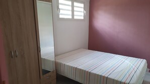 A bedroom with a bed, a wooden wardrobe, a window, and a pink wall. - Between Earth and sea (Le Lamentin)