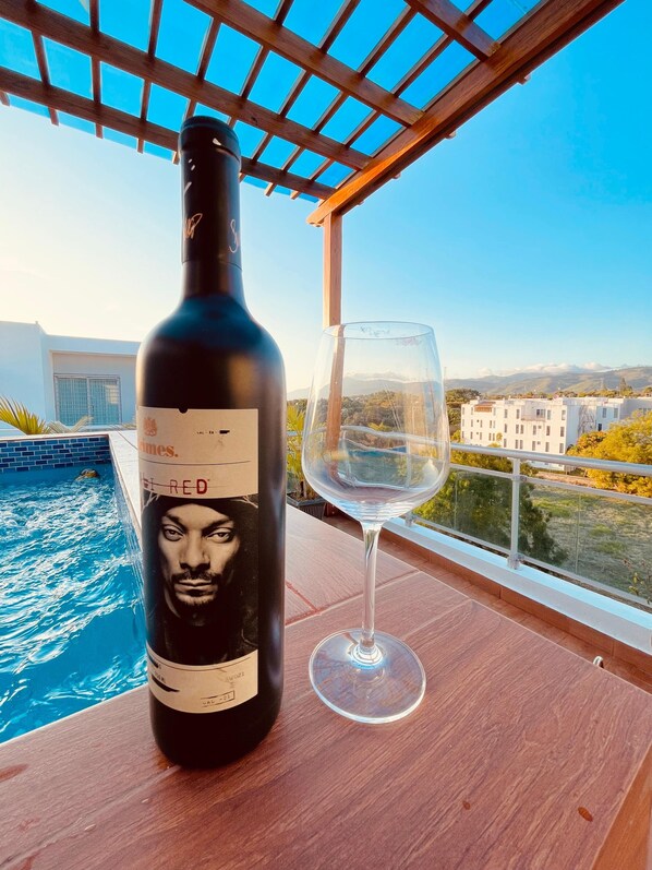 A bottle of red wine and an empty glass on a wooden table by a pool with a view of buildings and hills.