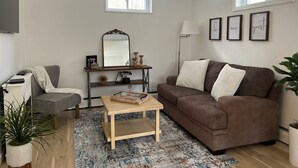 A living room with a brown sofa, a wooden coffee table, a small side table, a floor lamp, a mirror, and a potted plant.