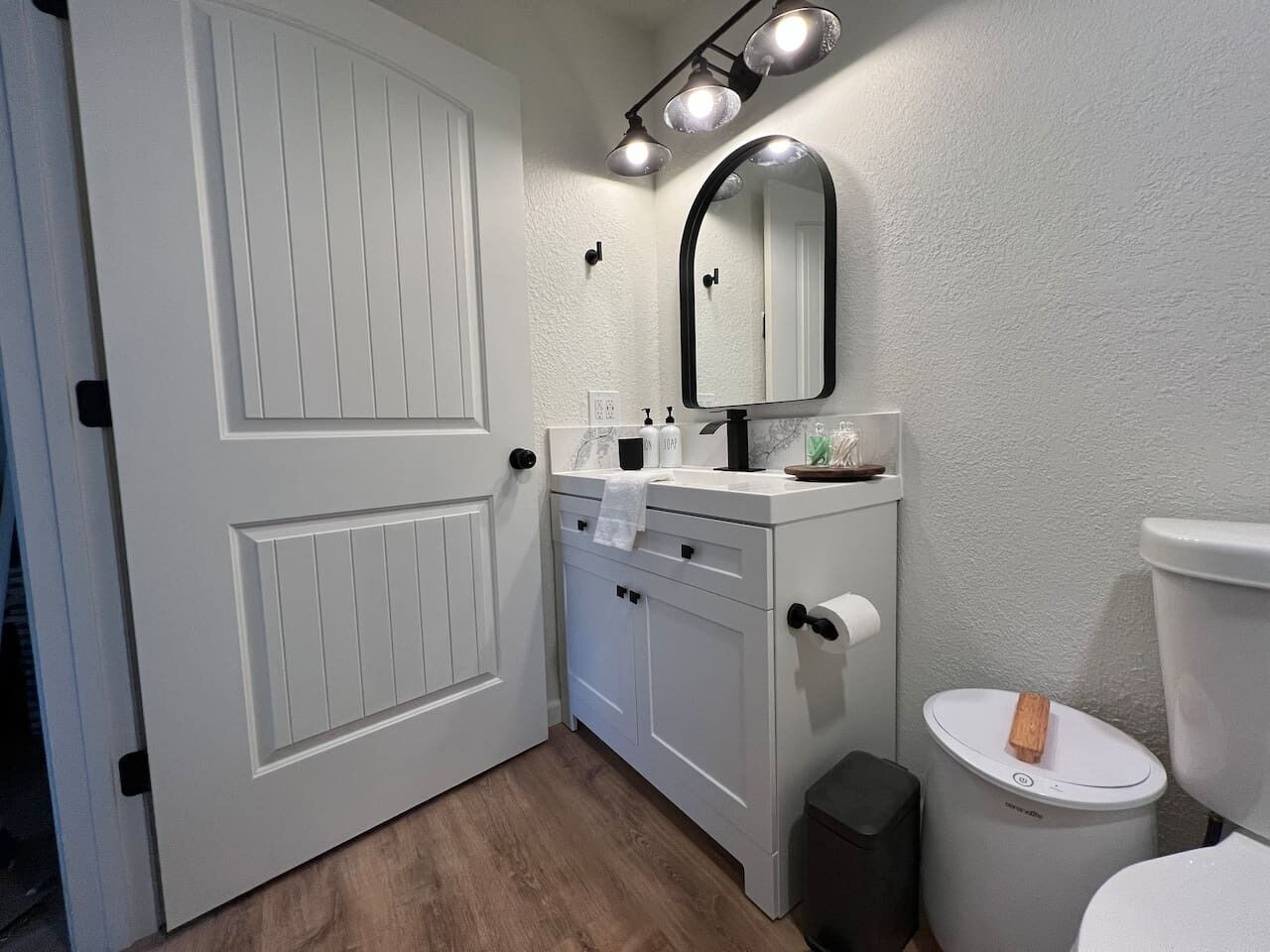 A bathroom with a white door, a vanity with a mirror, a toilet, and a trash can.