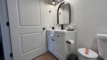 A bathroom with a white door, a vanity with a mirror, a toilet, and a trash can.