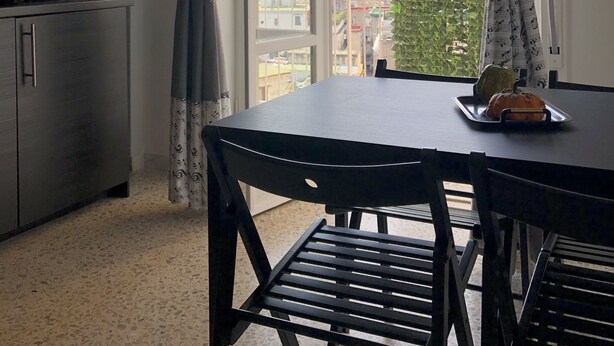 Uma sala de jantar com mesa e cadeiras, uma televisão na parede e vista para a cidade através de grandes janelas.