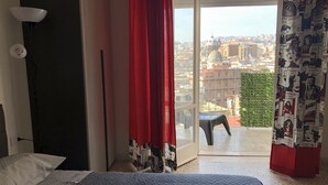 Quarto de hotel com cama, escrivaninha, cadeira, janela com cortinas vermelhas e vista para a paisagem urbana.