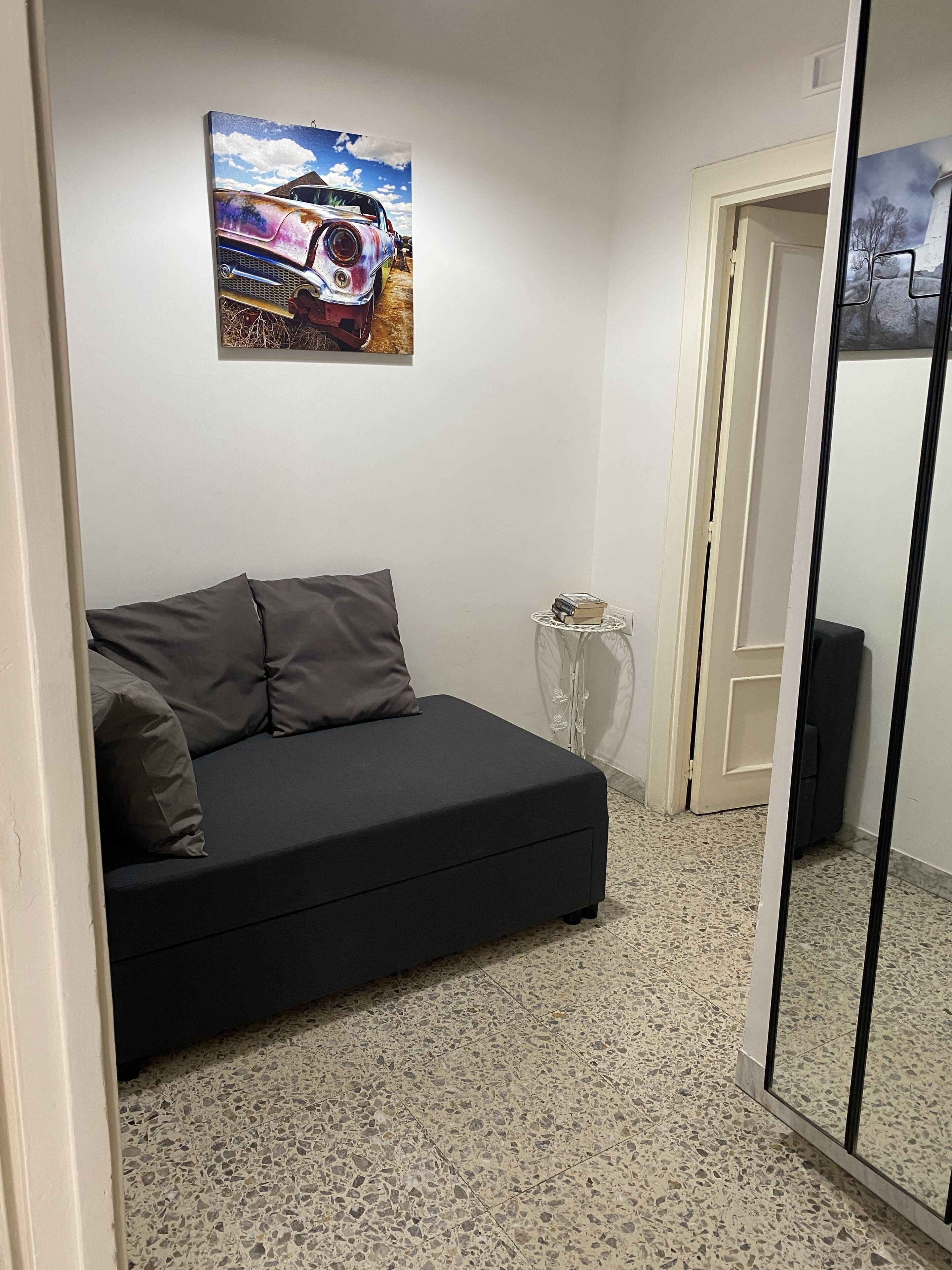 A room with a black sofa, a small round table with a lamp, a painting of a car on the wall, and a door leading to another room.