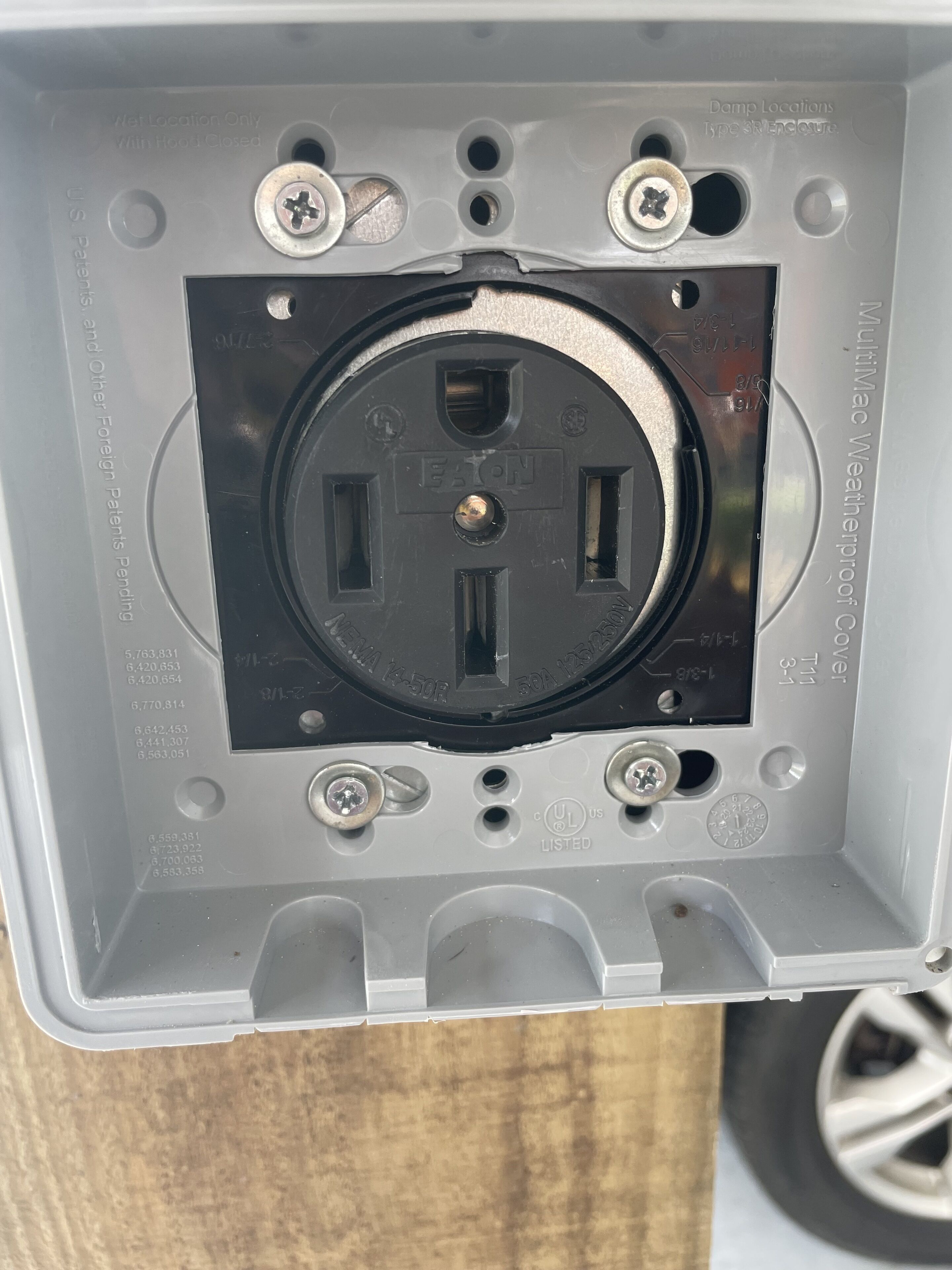 A power outlet with a black faceplate, surrounded by a metallic box with screw holes and a UL listed label.
