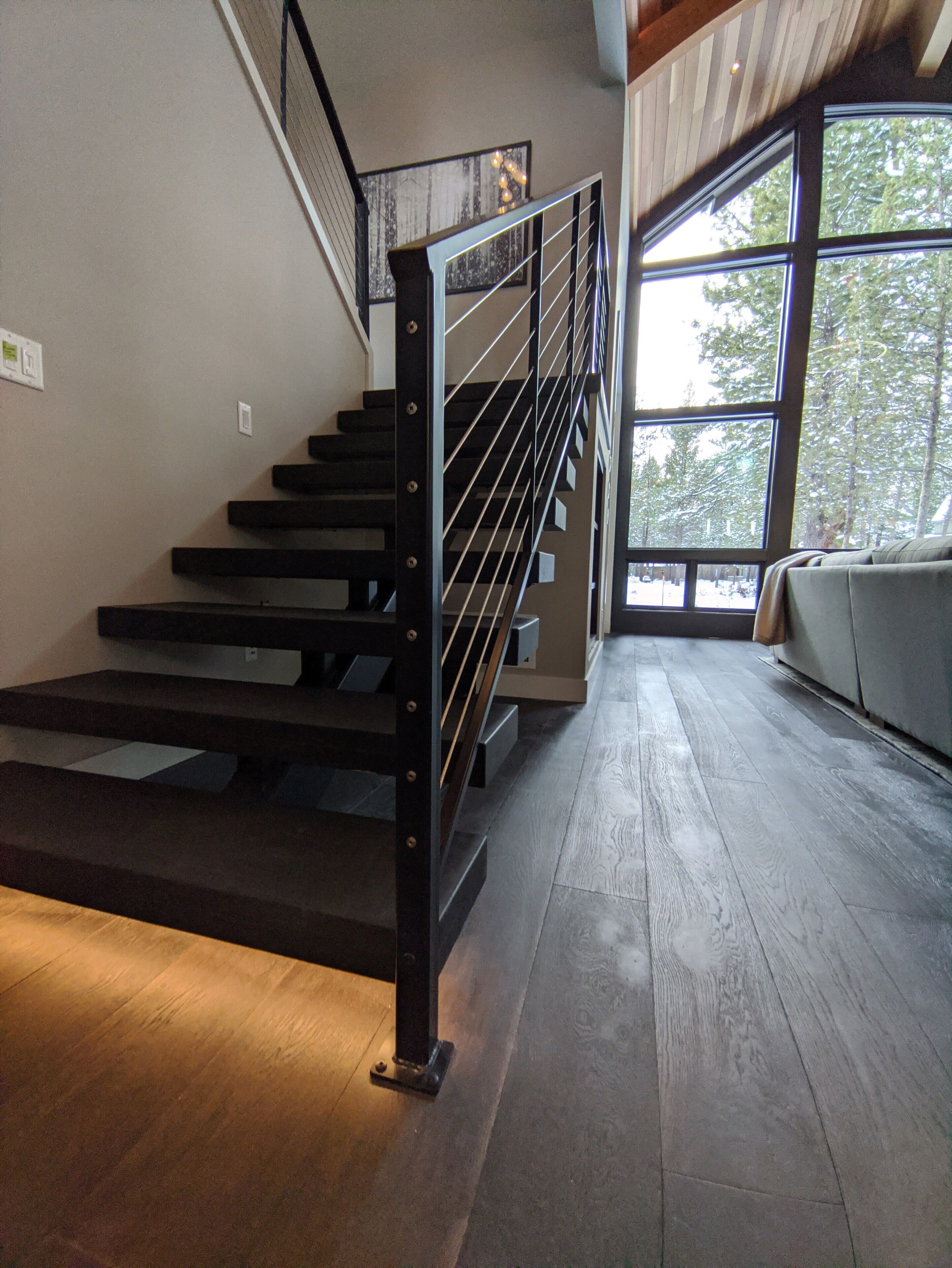 Un escalier avec une rampe en métal, des marches en bois et une vue sur les arbres à travers de grandes fenêtres.