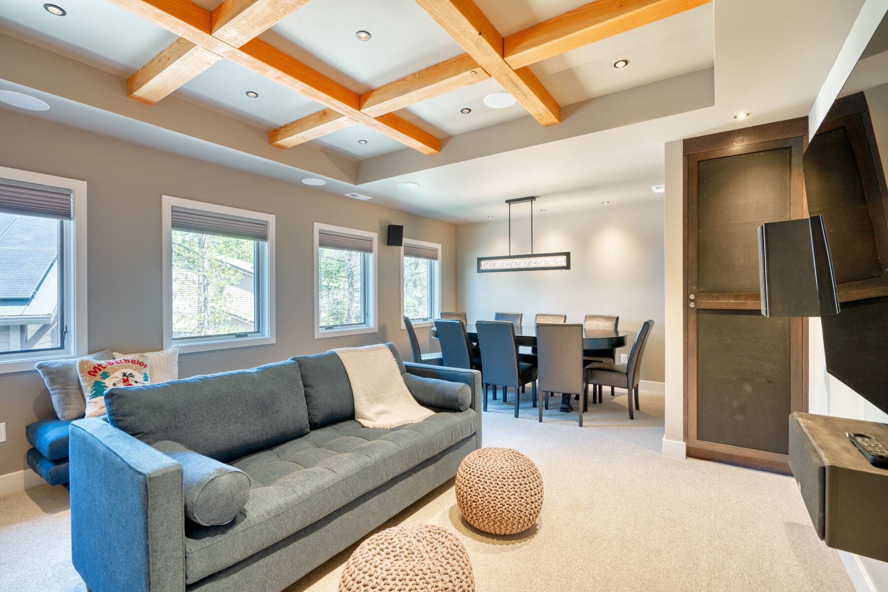 Un salon moderne avec un canapé gris, des poufs ronds, un coin repas comprenant une table et des chaises, et un téléviseur fixé sur un meuble en bois.
