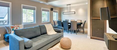 Un salon moderne avec un canapé gris, des poufs ronds, un coin repas comprenant une table et des chaises, et un téléviseur fixé sur un meuble en bois.